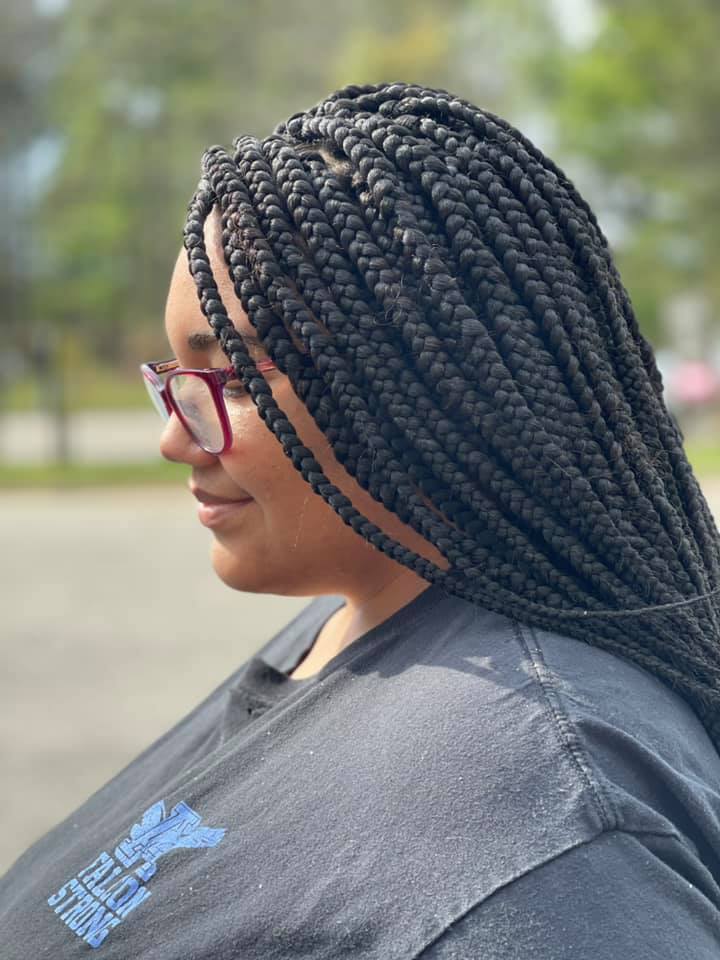 Side view of a woman wearing long, neatly parted box braids that fall over her shoulder. She is wearing red-framed glasses and a dark gray shirt with a light blue graphic, standing outdoors with a blurred background of trees and pavement.