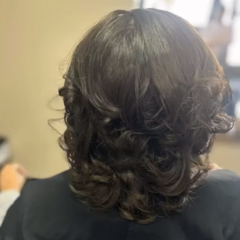 Back view of a person with shoulder-length, dark brown hair styled in soft, voluminous curls. The hair has a smooth, shiny finish with gentle waves and a natural, bouncy texture.